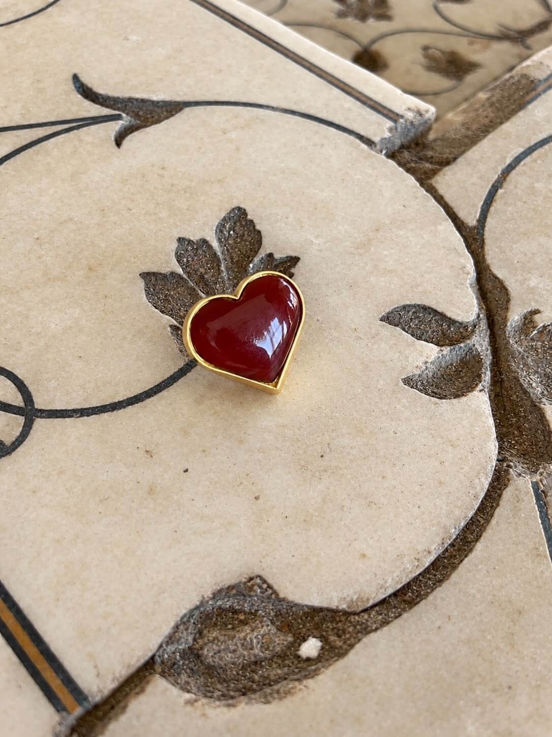 Heart Ring with a glossy deep red heart-shaped centre and gold-tone border, resting on floral-patterned stone.