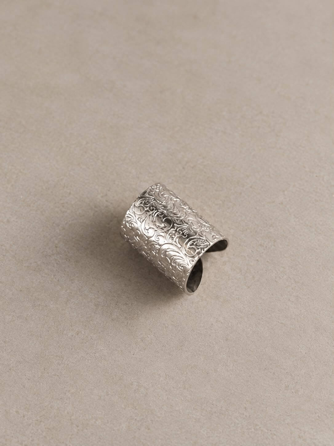 MEHRAB wide silver-tone ring with engraved floral scrollwork and an open-back band, displayed on beige.