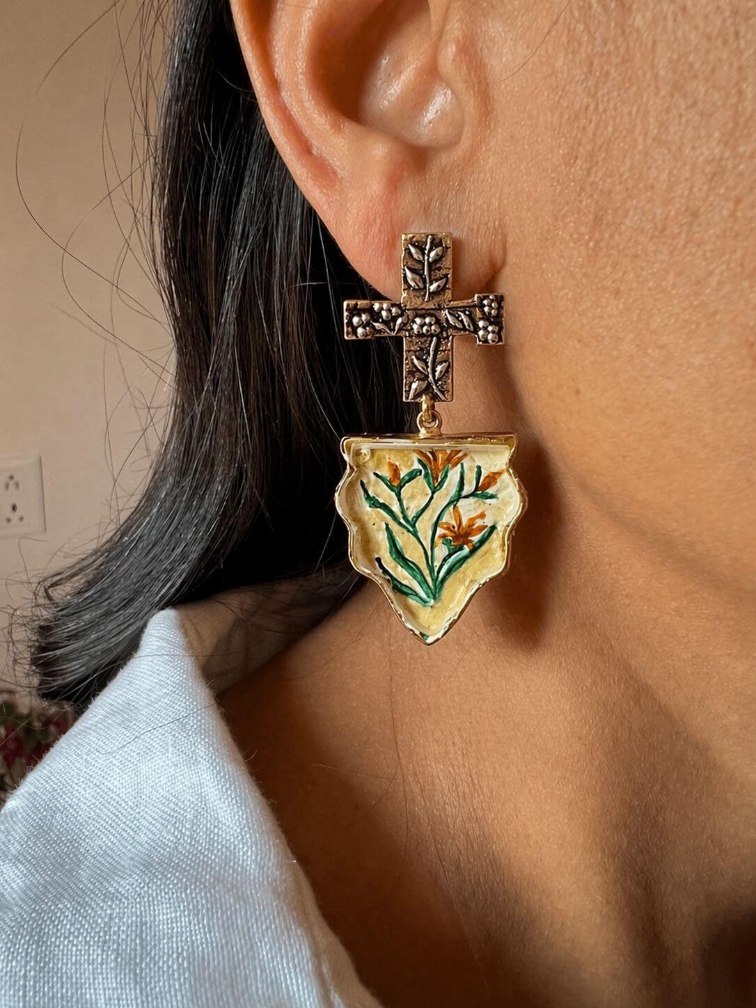 NAQASHE statement earring worn on an ear, showing an engraved cross-shaped stud and cream pendant with green and orange flowers.