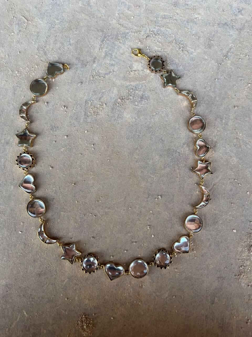 NOOR necklace with linked reflective hearts, stars, crescent moons and circles, laid flat on grey stone.