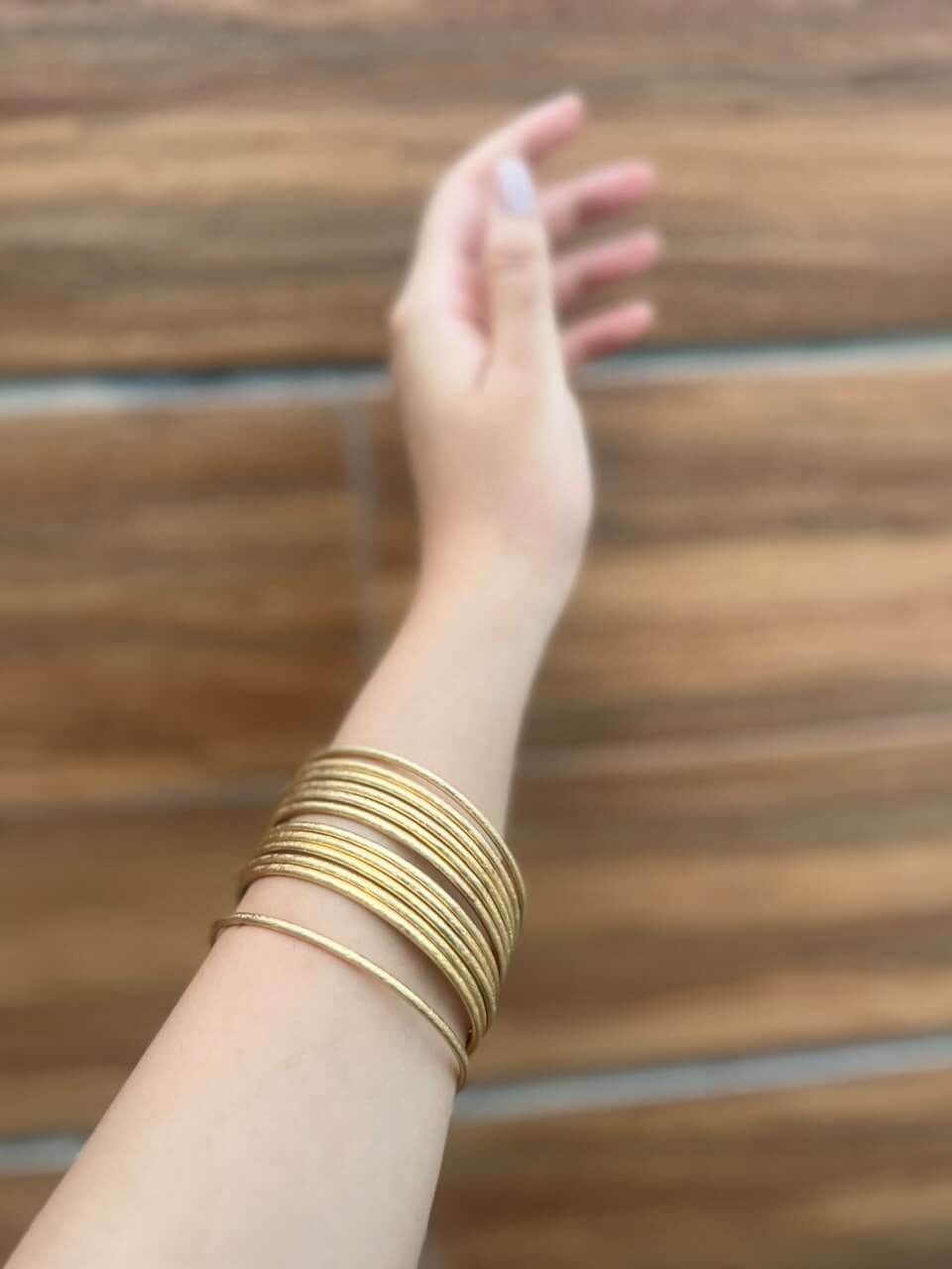 SONA CHURRI gold-tone bangles worn in a layered stack on a raised wrist against a wooden background