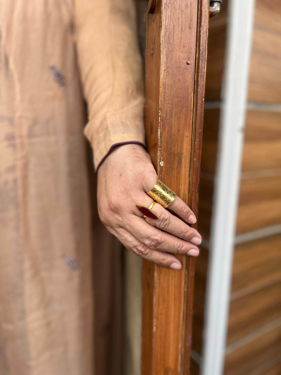 MEHRAB gold-tone engraved statement ring worn on a hand beside a red marquise ring against a wooden doorframe.